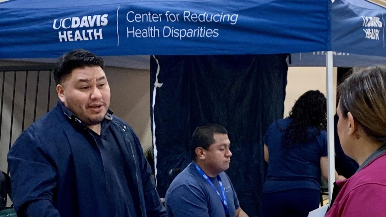 Indoor health fair with a blue UC Davis Health tent and table, with people interacting.